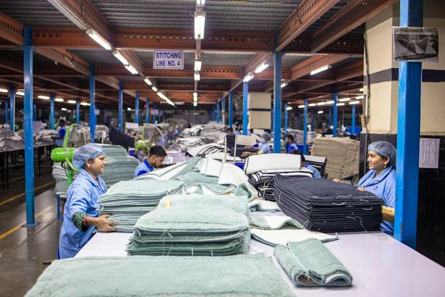 Workers handling stacks of textiles on an assembly table inside a large garment factory.